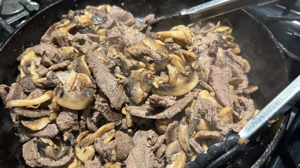 Beef and mushrooms folded back together in the skillet, tongs resting in the pan