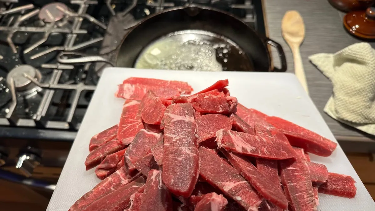 A board of sliced beef in the foreground, butter melting in a cast iron skillet on the gas range behind