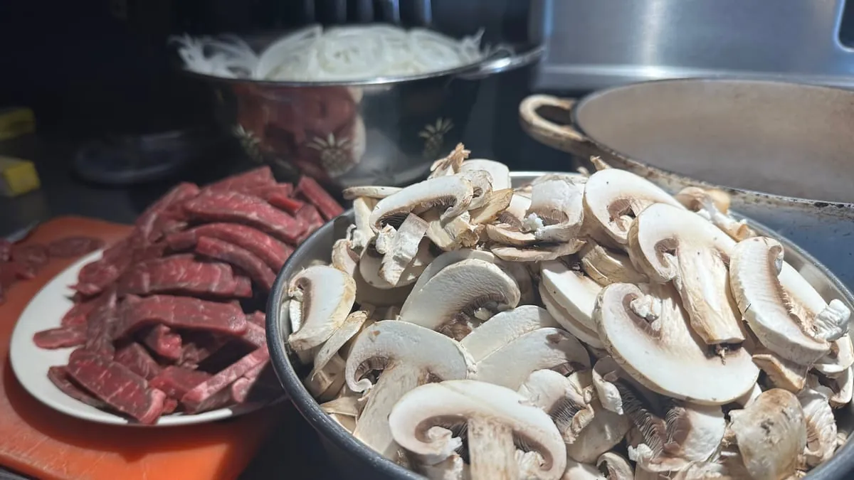 Sliced sirloin on a plate beside a bowl of sliced mushrooms, onions behind, in a dim kitchen