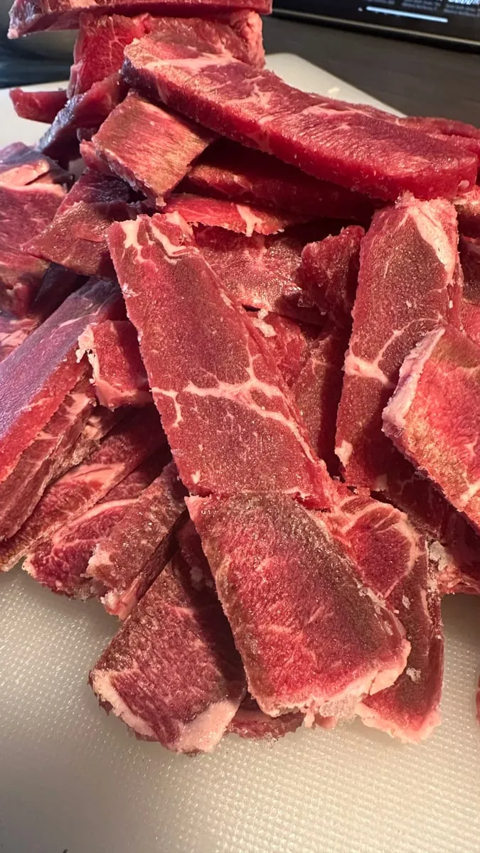 Strips of top sirloin still frosty from the freezer, stacked on a white cutting board