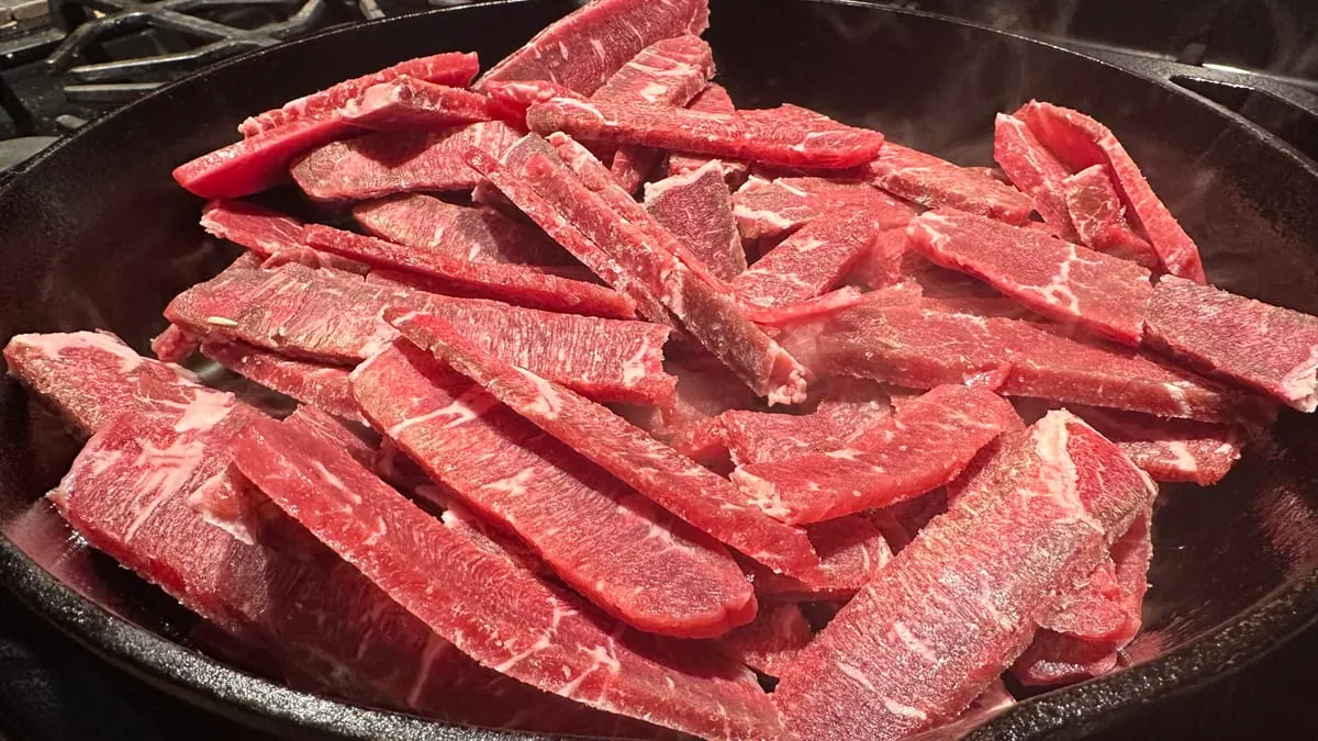 Raw strips of sirloin laid out in a single layer in a hot cast iron skillet